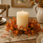 A lit cream pillar candle centered on a woven table runner, surrounded by orange and red berry branches with autumn leaves. The scene is set on a wooden dining table with plates, a ceramic pitcher, and a chair in a softly lit indoor setting.