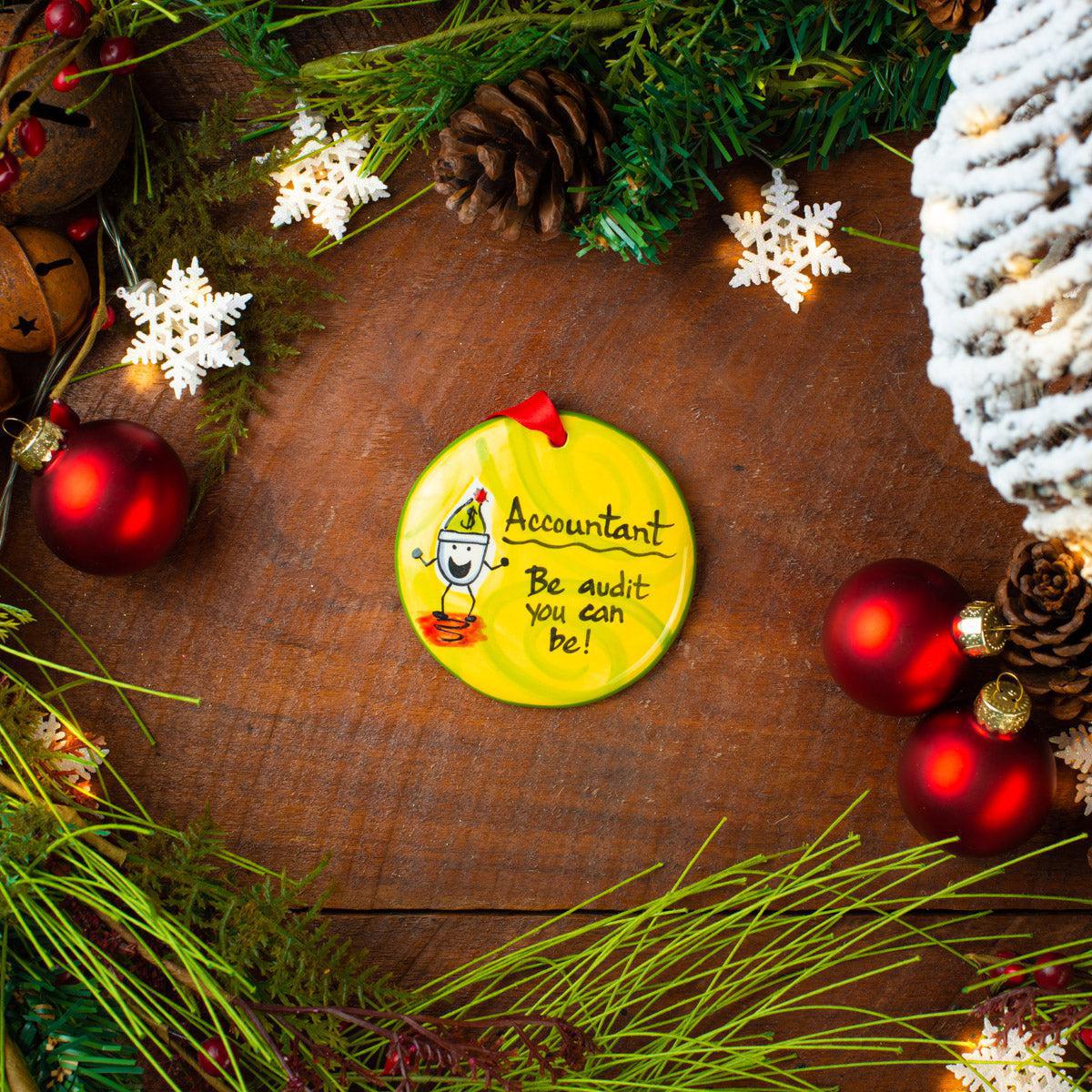 A ceramic Christmas ornament with a yellow background and a cartoon accountant design, placed on a festive decorated table with red ornaments and green foliage.