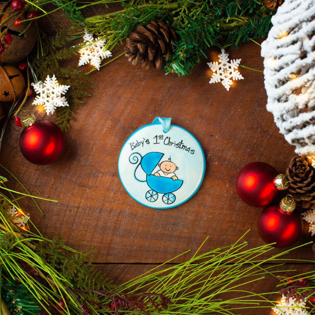 A round ceramic Christmas ornament with a hand-painted illustration of a baby in a stroller, labeled 'Baby's 1st Christmas'. The ornament is displayed on a wooden surface surrounded by festive decorations like pine cones, red ornaments, and green foliage.