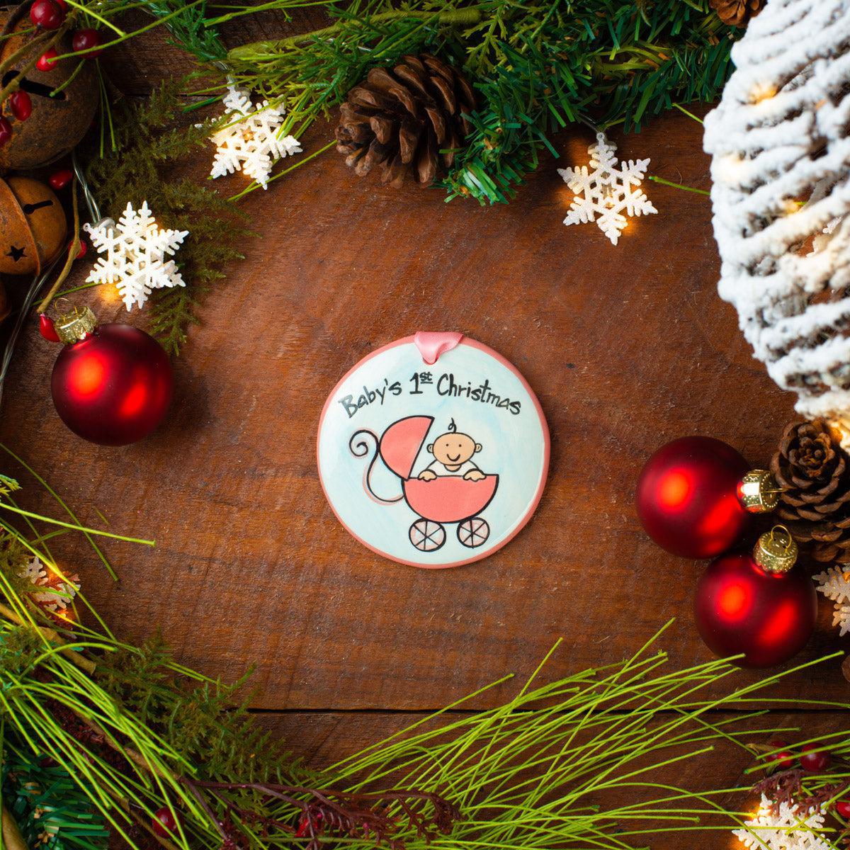 A round ceramic Christmas ornament with a hand-painted illustration of a baby in a stroller, labeled 'Baby's 1st Christmas'. The ornament is displayed on a wooden surface surrounded by festive decorations like pine cones, snowflakes, and red ornaments.