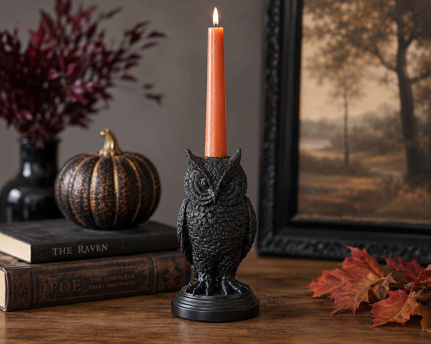 A black owl-shaped candle holder on a wooden table, holding a lit orange taper candle. The scene is styled with vintage books, a small pumpkin, red leaves, and a framed landscape print in the background.