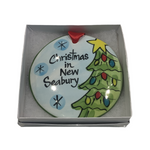 A flat, round ceramic Christmas ornament with a Christmas tree and the text 'Christmas in New Seabury'. The ornament is presented in a white gift box with a red ribbon.