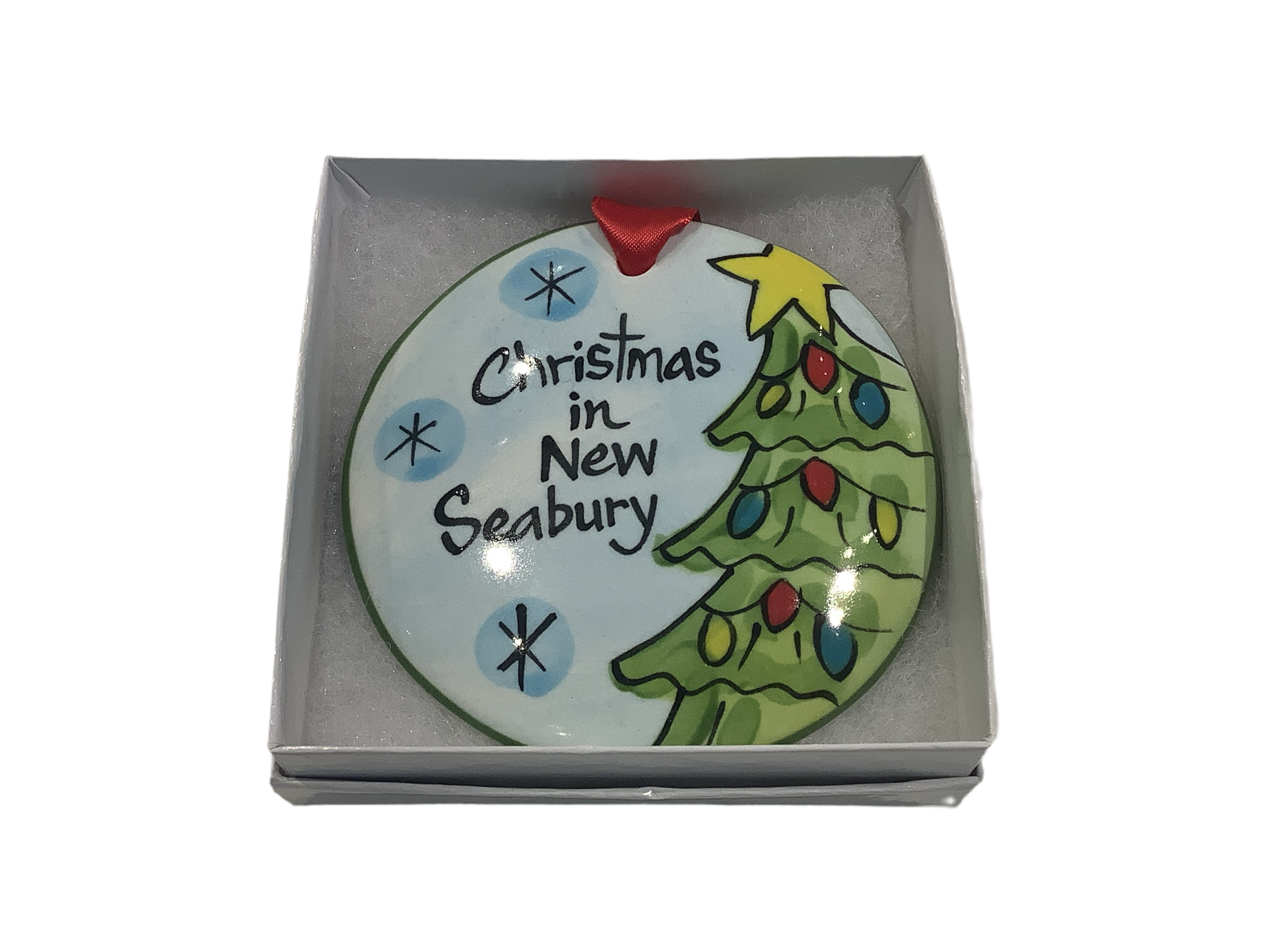 A flat, round ceramic Christmas ornament with a Christmas tree and the text 'Christmas in New Seabury'. The ornament is presented in a white gift box with a red ribbon.