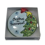 A flat, round ceramic ornament with a Christmas tree and the text 'Christmas in Popponesset' painted on it, packaged in a gift box.