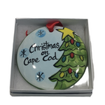 A flat, round ceramic Christmas ornament with a Christmas tree and the text 'Christmas on Cape Cod', presented in a gift box with a red ribbon.