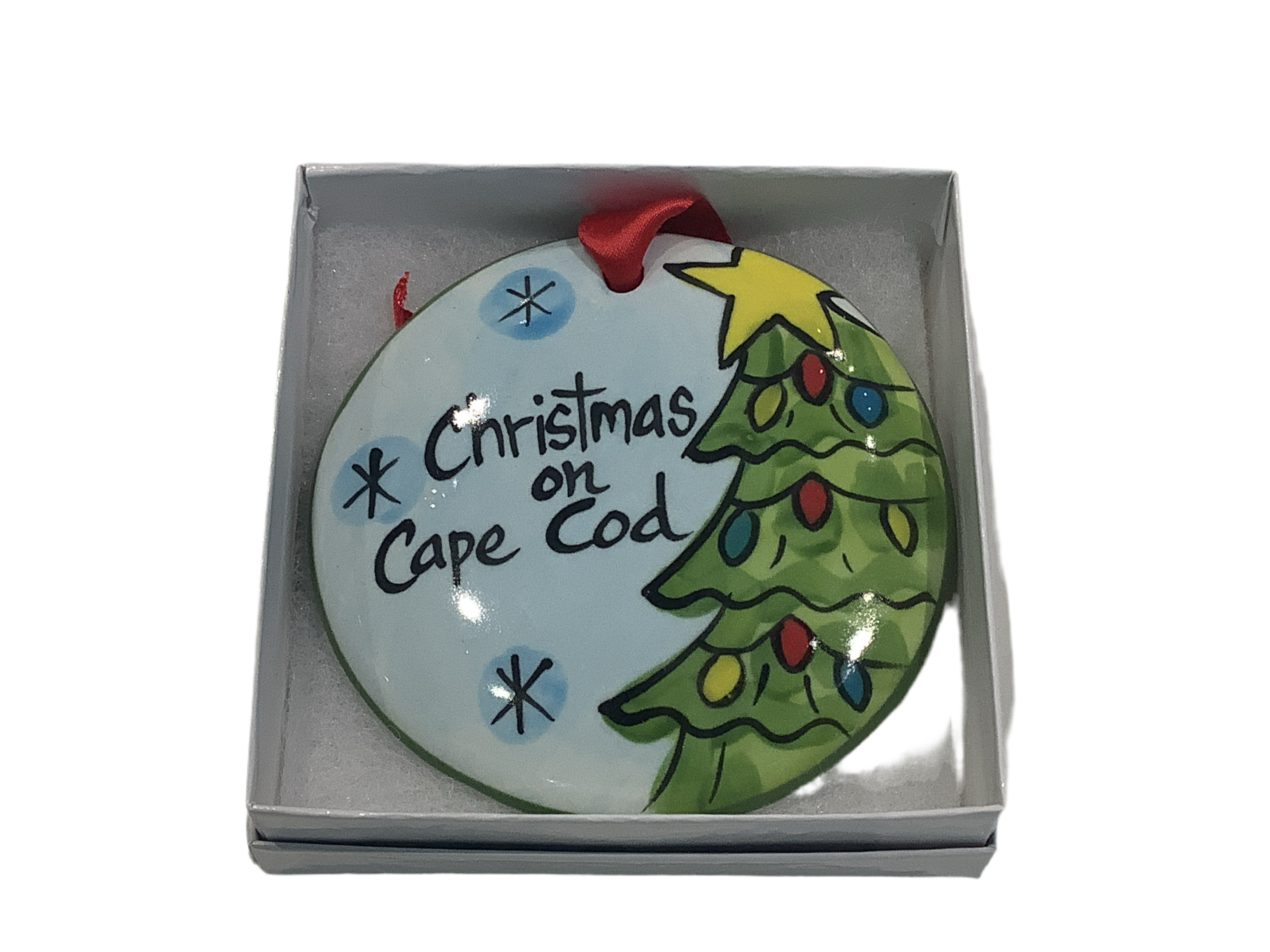 A flat, round ceramic Christmas ornament with a Christmas tree and the text 'Christmas on Cape Cod', presented in a gift box with a red ribbon.