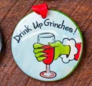 A flat, round ceramic ornament with a hand holding a red drink glass and the text 'Drink Up Grinches!' written on it.