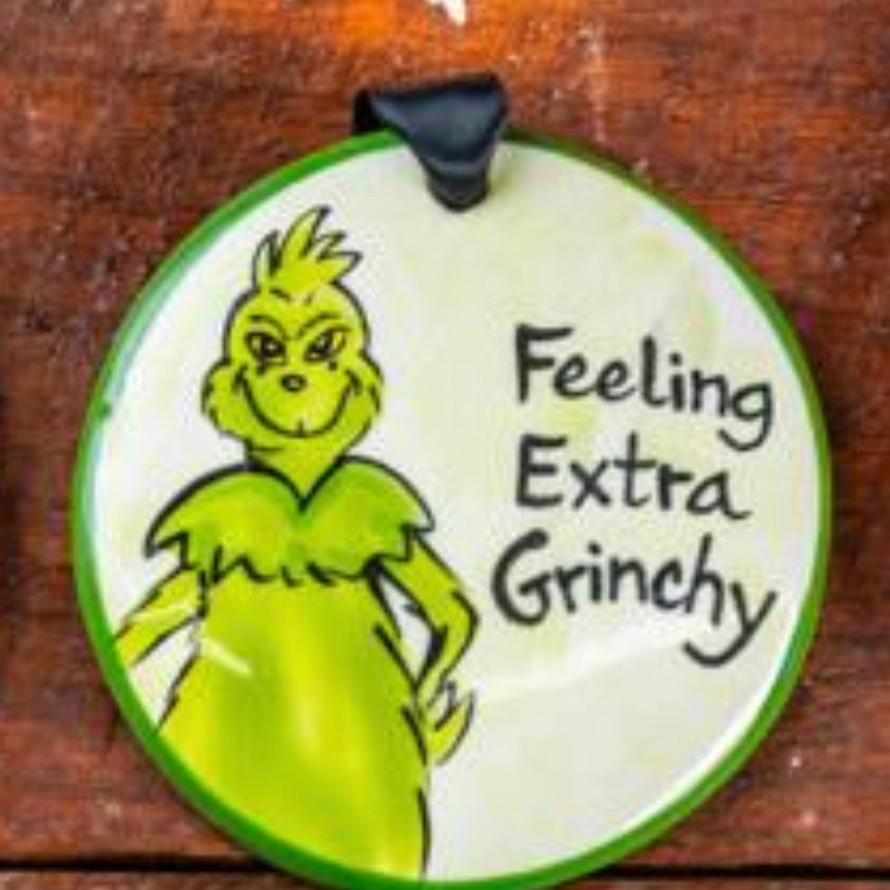 A round ceramic ornament with a hand-painted illustration of the Grinch and the text 'Feeling Extra Grinchy'.