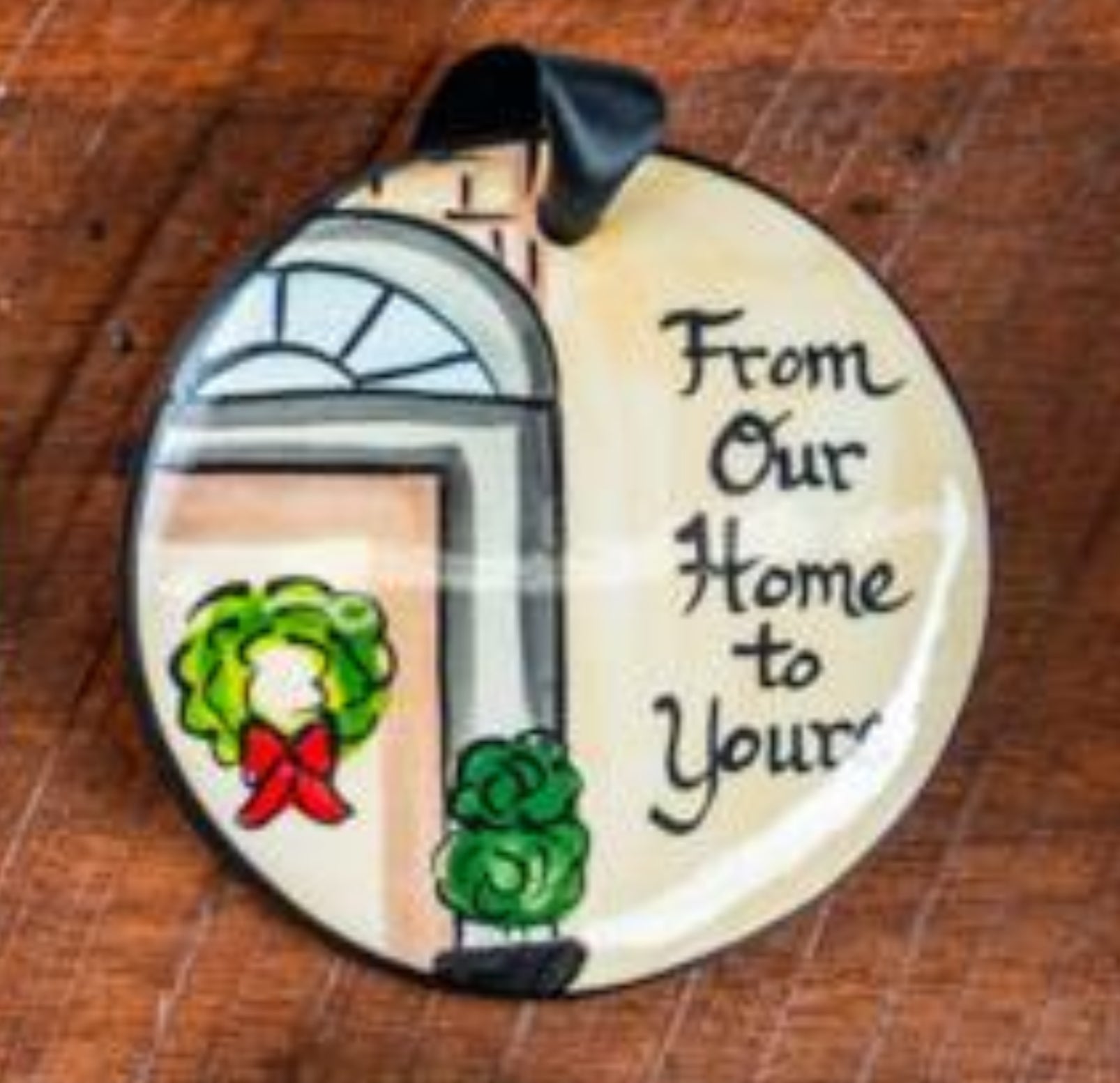A flat, round ceramic ornament with a Christmas theme, featuring a doorsteps scene, a wreath, and the message 'From Our Home to Your'.