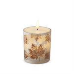 A lit taper candle in a clear glass jar decorated with metallic gold maple leaves, set against a plain white background.