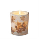 A lit taper candle in a clear glass container decorated with metallic gold maple leaves on a white background.