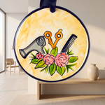 A round hanging ornament with a navy blue border and a yellow watercolor-style background, featuring the word “Hairdresser” above illustrated salon tools and pink flowers. The ornament is displayed against a dark textured backdrop, with a small gray vase below it.