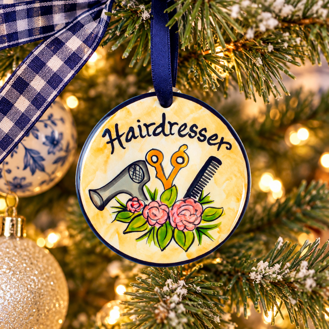 A round hanging ornament with a navy blue border and a yellow watercolor-style background, featuring the word “Hairdresser” above illustrated salon tools and pink flowers. The ornament is displayed against a dark textured backdrop, with a small gray vase below it.