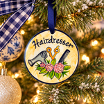 A round hanging ornament with a navy blue border and a yellow watercolor-style background, featuring the word “Hairdresser” above illustrated salon tools and pink flowers. The ornament is displayed against a dark textured backdrop, with a small gray vase below it.