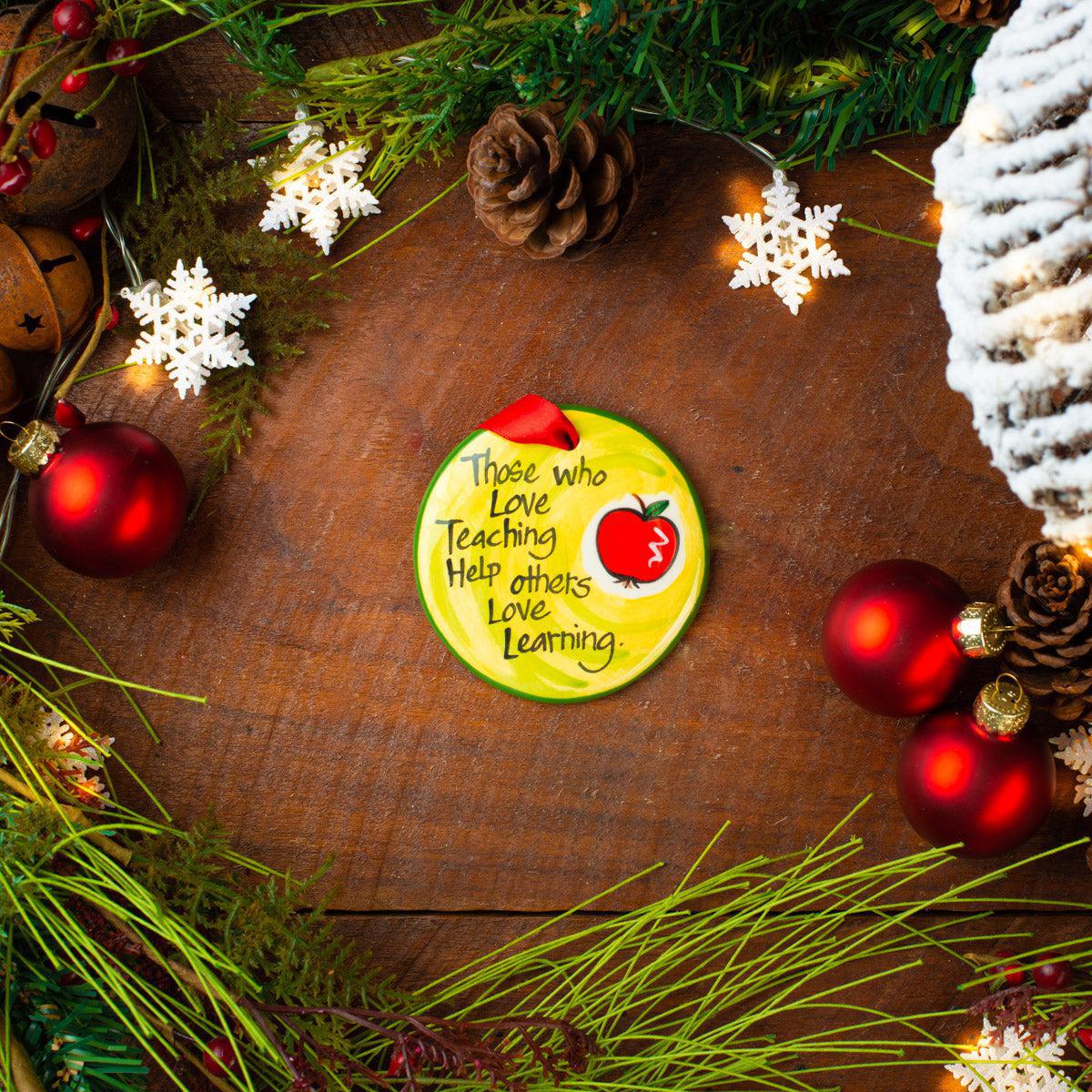 A circular ceramic ornament with a flat back, hand-painted with a green background, a red apple, and white text that reads 'Those who love teaching love learning.' The ornament is displayed on a wooden surface surrounded by holiday decorations including red bulbs, pine cones, and white snowflake decorations. Hand-painted ceramic teacher Christmas ornament with red apple and Those Who Love Teaching Help Others Love Learning quote, made in USA.