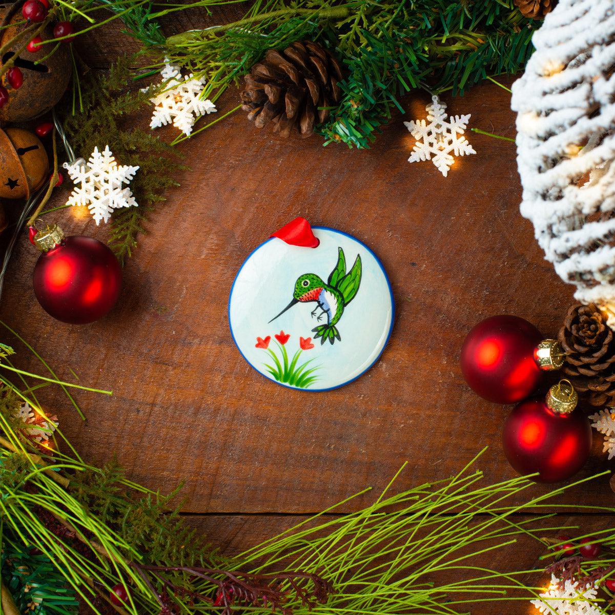 A ceramic hummingbird ornament painted in green with a red flower, displayed on a wooden surface surrounded by festive Christmas decorations including red bulbs, pine cones, and white snowflake ornaments.