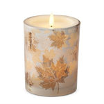 A lit taper candle in a cylindrical glass holder decorated with metallic gold maple leaves on a light beige background, set against a plain white studio backdrop.