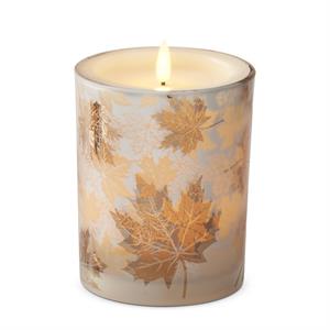 A lit taper candle in a cylindrical glass holder decorated with metallic gold maple leaves on a light beige background, set against a plain white studio backdrop.
