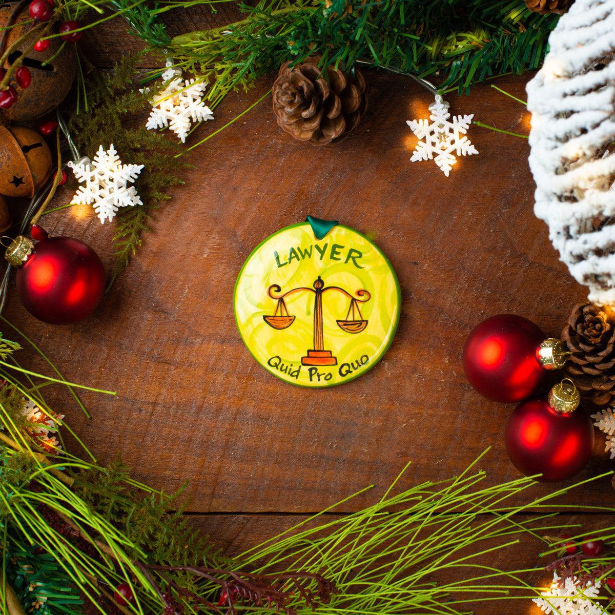 A ceramic Christmas ornament with a green and yellow design, featuring the word 'LAWYER' and the Latin phrase 'Justitia Pro Gloria', placed among holiday decorations on a wooden surface. Hand-painted ceramic lawyer Christmas ornament with scales of justice, Quid Pro Quo sentiment, and yellow green design, made in USA.