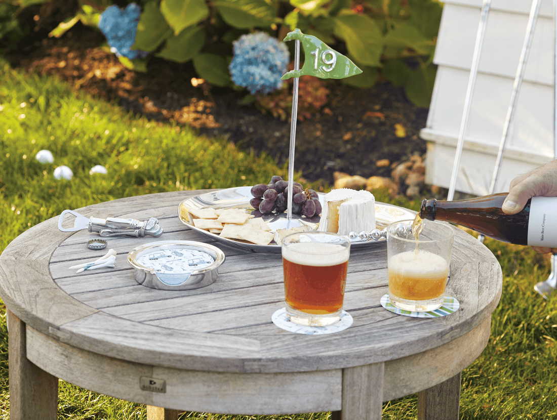Outdoor setting with a wooden table, drinks, snacks, and a person pouring beer.