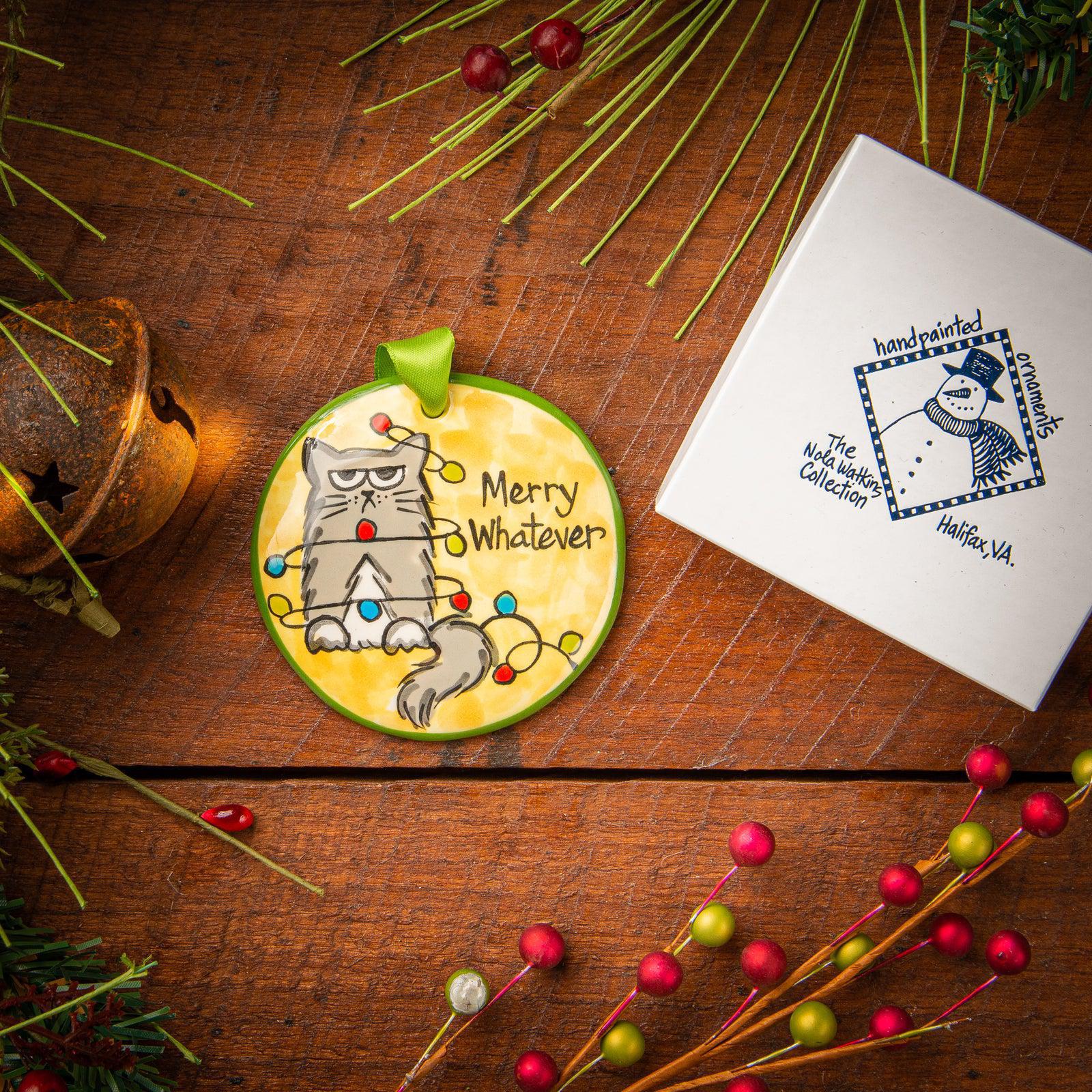 A ceramic Christmas ornament with a cartoon cat design, placed on a wooden surface with holiday decorations around it. A gift box is also visible next to the ornament.
