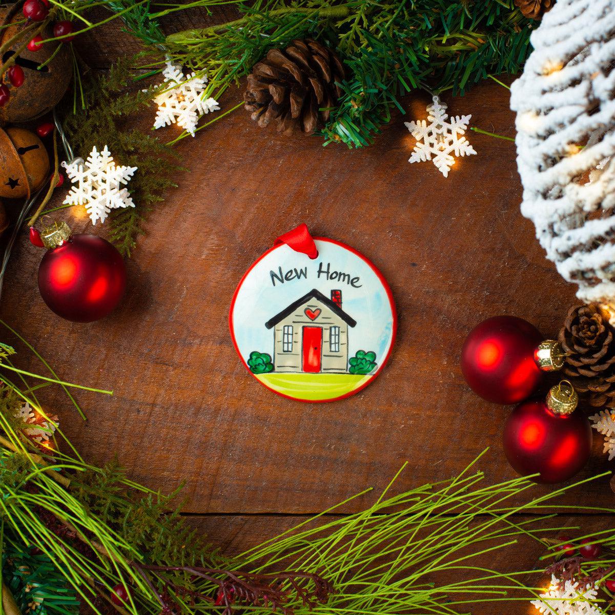 A ceramic holiday ornament with a 'New Home' motif, displayed on a wooden surface surrounded by festive decorations like pine cones and red baubles.