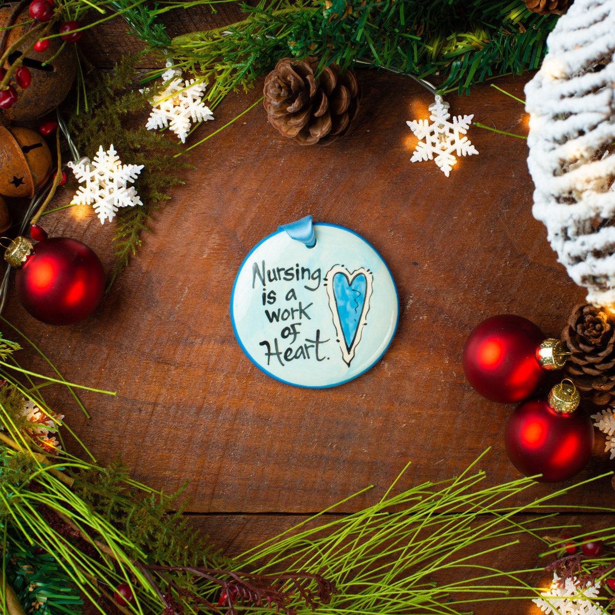 A round ceramic ornament with the text 'Nursing is a Work of Heart' painted on it, displayed among Christmas decorations on a wooden background. Hand painted in USA