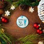 A round ceramic ornament with the text 'Nursing is a Work of Heart' painted on it, displayed among Christmas decorations on a wooden background. Hand painted in USA
