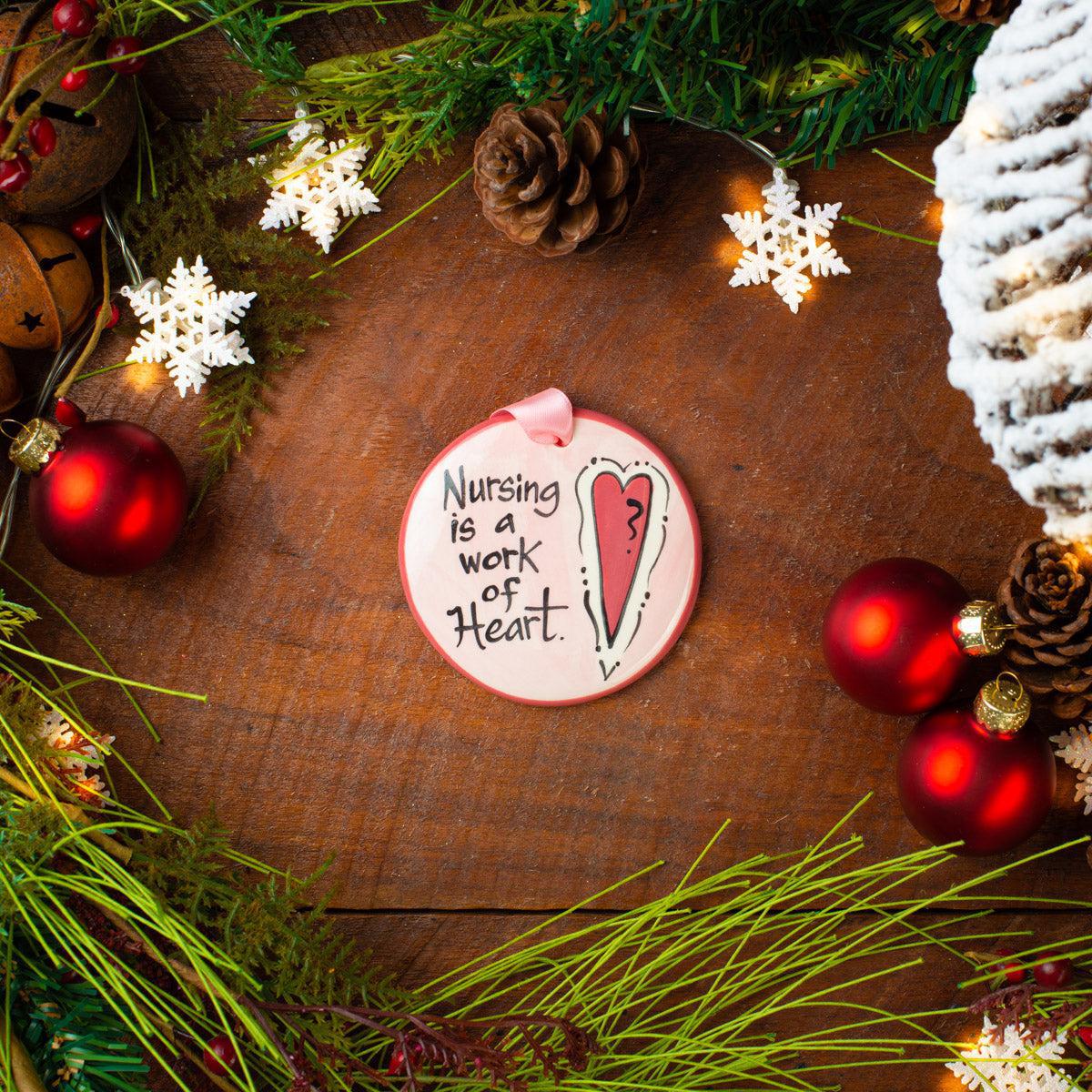 A round ceramic ornament with the text 'Nursing is a Work of Heart' painted on it, displayed among Christmas decorations on a wooden background. Hand painted  in USA