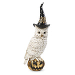 A white owl figurine wearing a black witch hat with gold accents and standing on a carved jack-o'-lantern pumpkin, photographed against a plain white background.