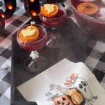 Halloween-themed cocktail setup with two pink drinks in coupe glasses garnished with orange slices and a heart, a bowl of punch, black marquee letter decorations, a black cauldron, spiderwebs, and a white napkin printed with pumpkins, ghosts, and the phrase 'WHAT'S YOUR poison?'.