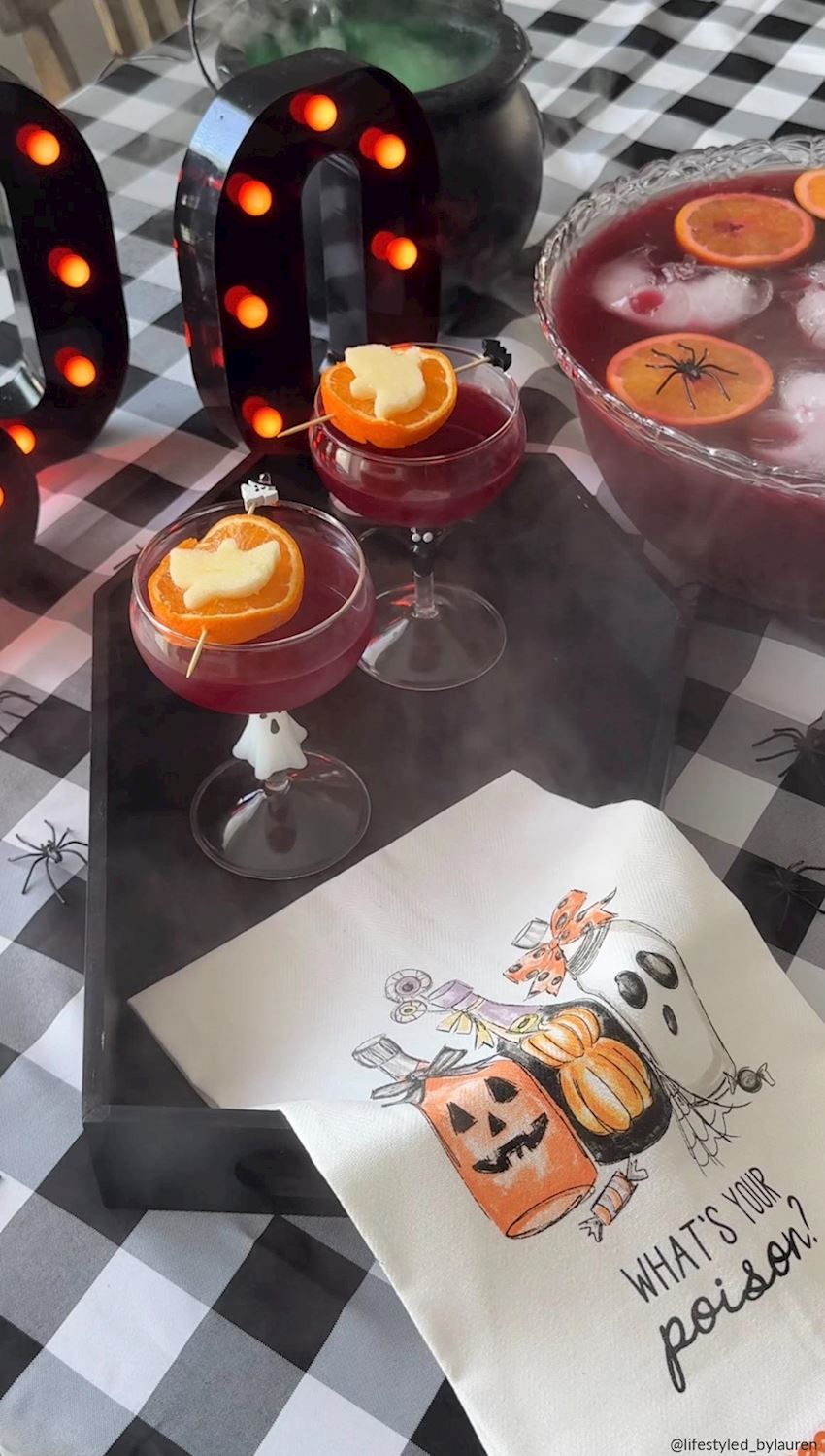 Halloween-themed cocktail setup with two pink drinks in coupe glasses garnished with orange slices and a heart, a bowl of punch, black marquee letter decorations, a black cauldron, spiderwebs, and a white napkin printed with pumpkins, ghosts, and the phrase 'WHAT'S YOUR poison?'.