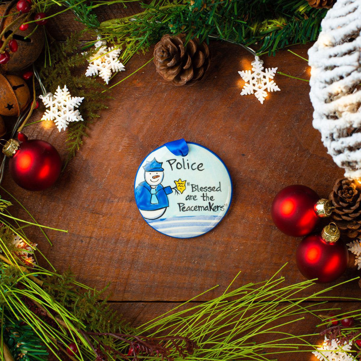 A circular ceramic ornament with a cartoon depiction of a police officer and the text 'Police Blessed are the Peacemakers'. The ornament is surrounded by Christmas decorations on a wooden background.