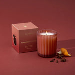 A lit Thymes Pomander Clove scented candle in a ribbed glass jar sits next to its dusty rose packaging box on a matching dusty rose background, styled with star anise and a citrus peel slice.