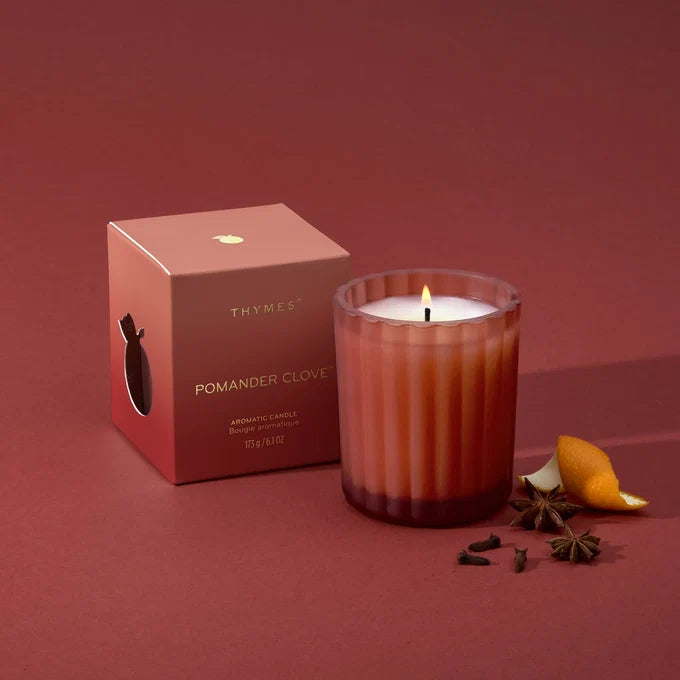 A lit Thymes Pomander Clove scented candle in a ribbed glass jar sits next to its dusty rose packaging box on a matching dusty rose background, styled with star anise and a citrus peel slice.