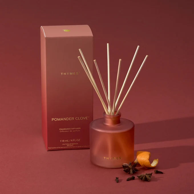 Thymès Pomander Clove reed diffuser set with a frosted glass bottle, natural reed sticks, and its matching pink packaging box on a matching dusty rose background, styled with orange peel and spices.