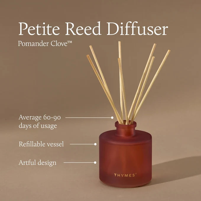 A reed diffuser bottle in a deep rose-red tone with natural reed sticks, shown on a warm beige background with text reading 'Petite Reed Diffuser' and 'Pomander Clove.' Callout lines mention average 60-90 days of usage, refillable vessel, and artful design.
