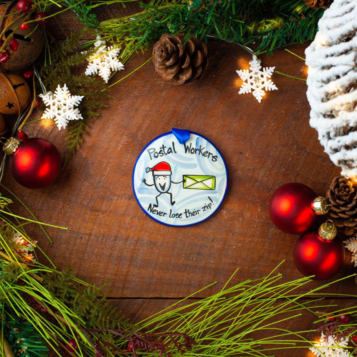 A ceramic ornament with a flat, disk shape featuring a illustration of a mailman and the text 'Postal Worker', surrounded by holiday decorations on a wooden background.