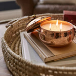 Copper candle holder with three candles on a book, placed on a woven tray.