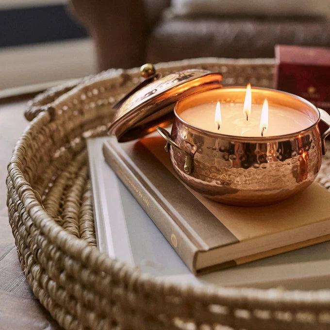 Copper candle holder with three candles on a book, placed on a woven tray.
